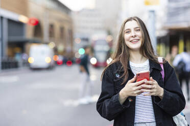 Happy young woman standing with smart phone in city - WPEF08676