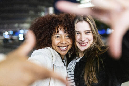 Smiling friends looking through finger frame at night - WPEF08697