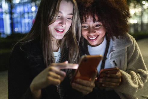 Smiling young women using smart phones at night - WPEF08705
