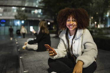 Glückliche junge Frau sitzt mit Smartphone in der Stadt bei Nacht - WPEF08718