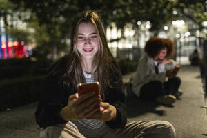 Lächelnde schöne Frau, die nachts ein Mobiltelefon benutzt - WPEF08719