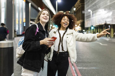 Happy young woman hailing ride with friend holding smart phone on street at night - WPEF08727
