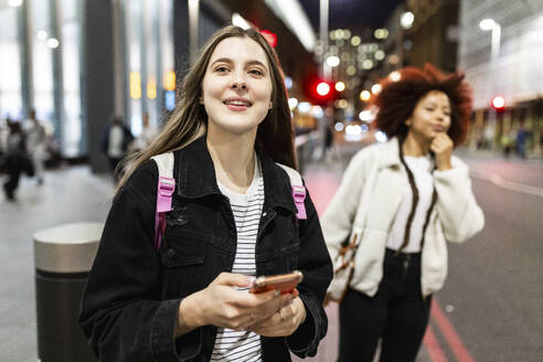 Smiling woman holding smart phone with friend standing on street - WPEF08729