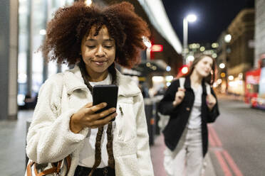 Lächelnde junge Frau mit Afrofrisur, die auf der Straße ein Mobiltelefon benutzt - WPEF08732