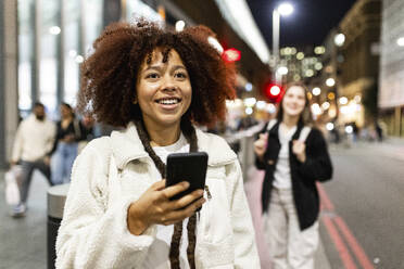 Happy woman holding smart phone and standing on street - WPEF08733