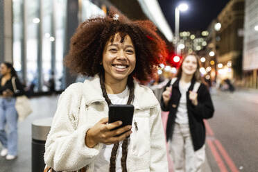 Happy young woman holding smart phone and standing on street - WPEF08734