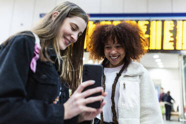 Smiling woman showing smart phone to friend at railroad station - WPEF08739