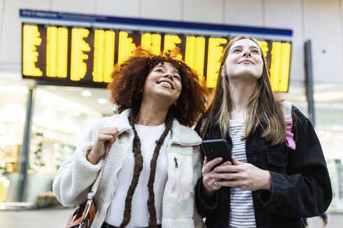 Smiling woman holding smart phone and standing with friend at railroad station - WPEF08742