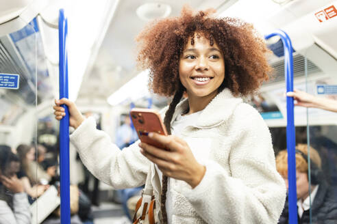Smiling woman standing with smart phone in subway train - WPEF08748