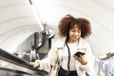 Lächelnde Frau, die auf einer U-Bahn-Rolltreppe ein Mobiltelefon benutzt - WPEF08753