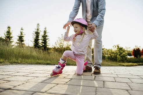 Father helping daughter wearing roller skates on footpath - NSTF00043