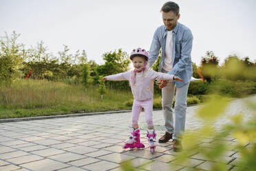 Happy girl learning to roller skate with father on footpath - NSTF00045