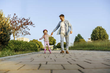 Father holding hand of daughter roller skating on footpath - NSTF00047