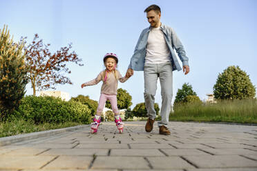 Happy girl learning to roller skate with father on footpath - NSTF00048