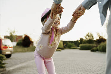 Fröhliches Mädchen, das sich an den Händen seines Vaters festhält und auf dem Fußweg Rollschuhlaufen lernt - NSTF00053