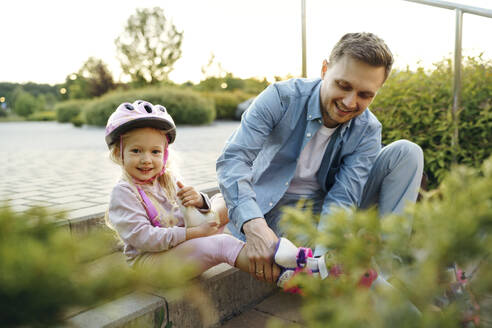 Smiling girl with father helping to wear roller skates sitting at park - NSTF00057