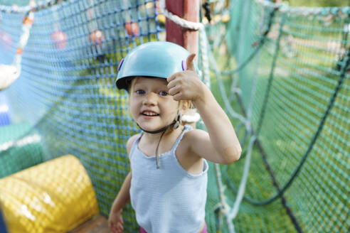 Cute girl wearing helmet and showing thumbs up gesture at rope park - NSTF00060