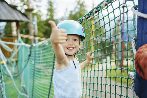 Excited smiling girl showing thumbs up gesture near net at rope park - NSTF00066