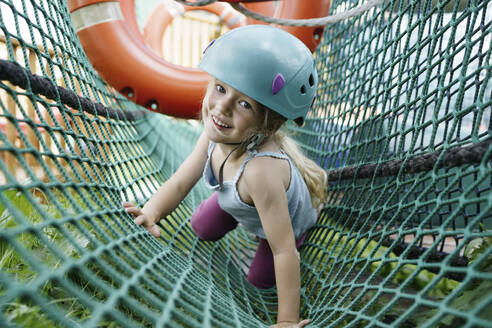 Happy girl wearing helmet and crawling on net at park - NSTF00069