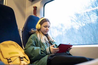 Mädchen mit drahtlosen Kopfhörern und Tablet-PC in der Nähe des Fensters im Zug sitzend - MDOF01937