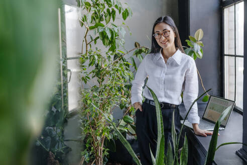 Smiling young businesswoman standing near laptop and plants in office - YTF02017