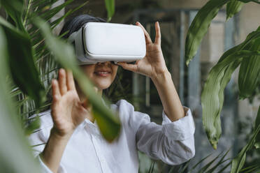 Businesswoman wearing virtual reality simulator and gesturing near plants - YTF02026