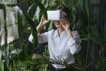 Smiling businesswoman wearing virtual reality simulator near plants - YTF02027