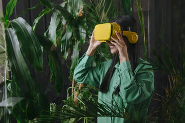 Happy businesswoman wearing yellow virtual reality simulator near plants - YTF02030