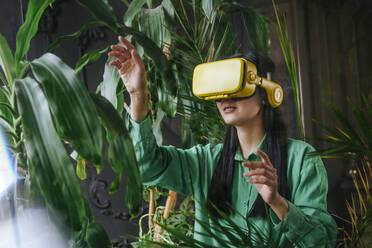 Businesswoman wearing yellow virtual reality simulator and gesturing near plants - YTF02032
