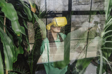 Smiling businesswoman wearing yellow virtual reality simulator seen through window - YTF02033