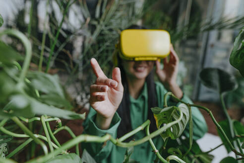 Junge Geschäftsfrau trägt gelben Virtual-Reality-Simulator und gestikuliert in der Nähe von Pflanzen - YTF02036