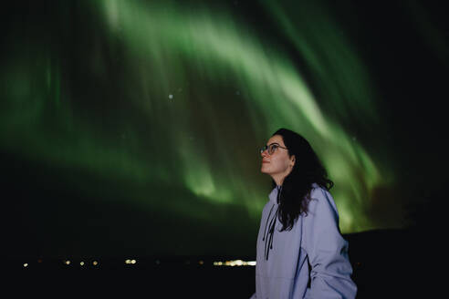 Side view woman stands under the mystical green glow of the Aurora Borealis during a cold night in Iceland. - ADSF53337