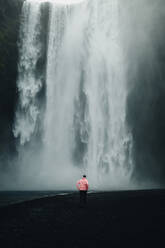 Back view of unrecognizable man in a pink jacket stands observing the majestic Skogafoss waterfall in Iceland, surrounded by mist and dark volcanic terrain. - ADSF53340