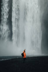 Back view of anonymous man in a vibrant orange jacket stands captivated by the Skogafoss waterfall in Iceland, with mist rising around the powerful cascade. - ADSF53341