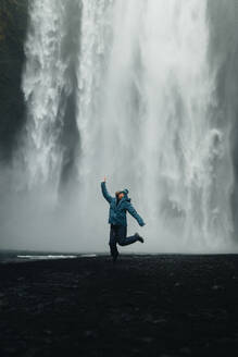 A female traveler expresses joy with a mid-air jump before the powerful backdrop of an Icelandic waterfall, capturing the thrill of exploration. - ADSF53342