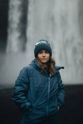 A female traveler wearing a warm beanie and blue jacket stands before a misty Icelandic waterfall, contemplating nature's serenity. - ADSF53343