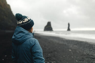 A woman traveler stands on the black sand beach at Reynisfjara, looking towards the iconic Reynisdrangar sea stacks off the coast of Iceland. - ADSF53346