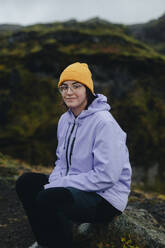 A young woman wearing a yellow beanie and purple jacket is sitting on a rock with a moss-covered Icelandic hill in the background. - ADSF53349