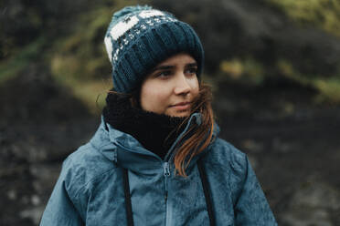 A young woman wearing a knitted beanie and a blue jacket stands thoughtful in the Icelandic wilderness. - ADSF53368