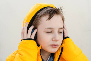 A contemplative young boy in a striking yellow jacket enjoys music on his headphones against a soft neutral background - ADSF53374