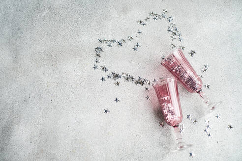 From above of two pink champagne glasses laid down with a trail of silver star-shaped confetti on a textured white surface - ADSF53377
