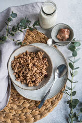 A nutritious granola bowl with a spoon, milk bottle, and eucalyptus decor on a woven placemat - ADSF53379