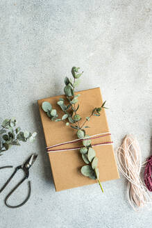 A completed gift wrapping with brown paper, eucalyptus leaves, and pink and white twine on a textured grey background, accompanied by scissors and extra twine - ADSF53384