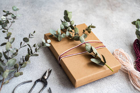 A beautifully wrapped gift in brown paper adorned with eucalyptus branches and twine, next to scissors and extra twine on a concrete background - ADSF53385