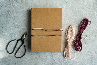 A simple yet elegant gift wrapped in brown paper with a pink and white twine, alongside scissors and additional twine on a textured grey background - ADSF53386