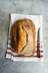 Top view of golden-brown rye sourdough bread on a white cloth with red stripes, next to a bread knife on a textured surface - ADSF53387