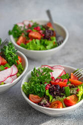 Freshly prepared vegetable salad featuring crisp lettuce, arugula, sliced radishes, cherry tomatoes, green onions, and pomegranate seeds in a white bowl on a textured grey surface - ADSF53389