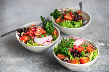 Three bowls of vibrant vegetable salad with lettuce, arugula, radishes, cherry tomatoes, green onions, and pomegranate seeds on a grey countertop - ADSF53390