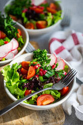 A close-up of a bowl of colorful vegetable salad with lettuce, arugula, radishes, cherry tomatoes, and pomegranate seeds on a wooden cutting board - ADSF53391