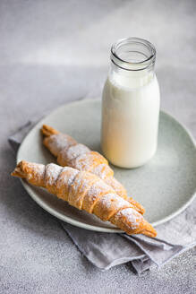 Zwei mit hausgemachter Marmelade gefüllte und mit Puderzucker bestäubte Törtchen, dazu eine Flasche Milch, auf einem Keramikteller über einer grauen Serviette und einer Arbeitsplatte - ADSF53394